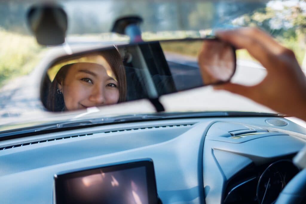 A person adjusting the rearview mirror in a car, smiling as their reflection is visible. Sunlight streams through the windshield, highlighting part of the car's interior. The road ahead is slightly out of focus.