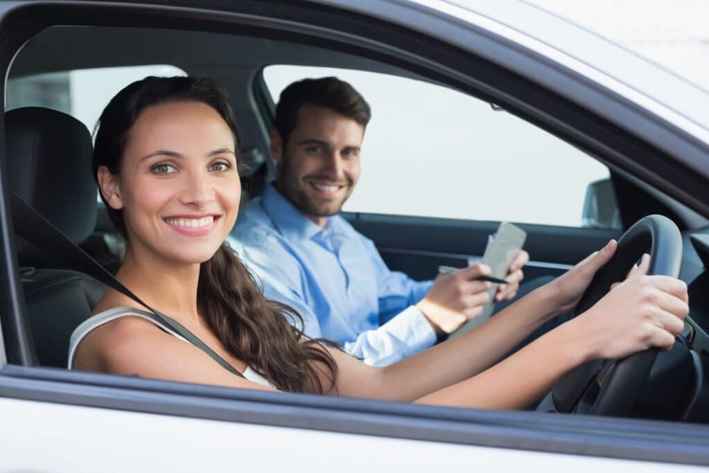 A woman is smiling as she takes her automatic driving lessons in Bristol, with a man beside her in the passenger seat, also smiling and holding a smartphone. The interior of the car is bright and modern.