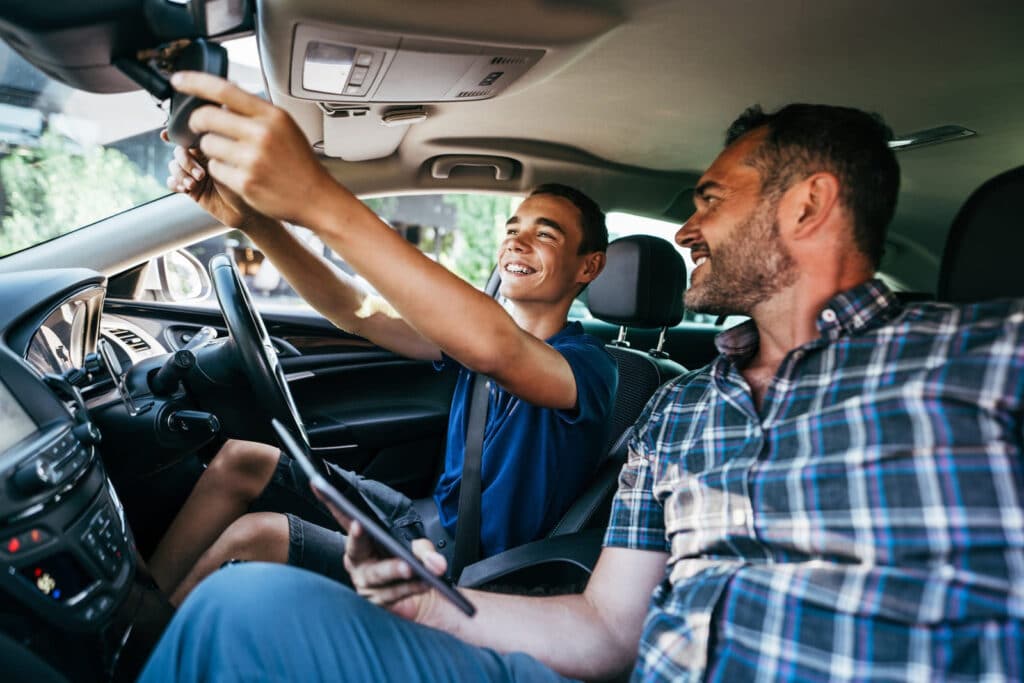 A smiling teenage boy is adjusting the rearview mirror in a car while an adult man, sitting in the front passenger seat, looks at him proudly. Both appear to be sharing a joyful moment inside a parked vehicle.