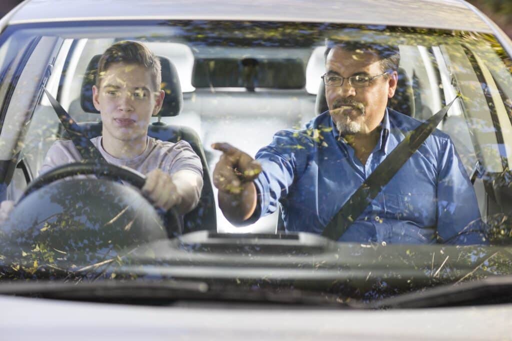 A young person driving a car with an older person in the passenger seat. The passenger is pointing ahead, possibly giving instructions. Sunlight and reflections are visible on the windshield.