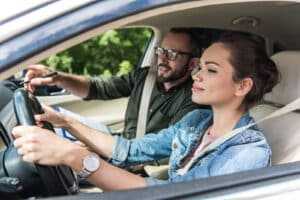 A woman is driving a car with a man sitting beside her, pointing at the dashboard. She looks focused, while he appears to be giving her instructions. Both are wearing seatbelts and the road is visible through the windshield.