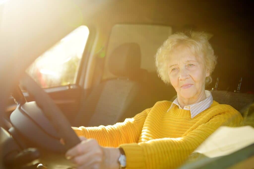 An elderly person with short white hair is sitting in the driver's seat of a car, wearing a yellow sweater. Sunlight is streaming through the window, creating a warm and bright atmosphere.