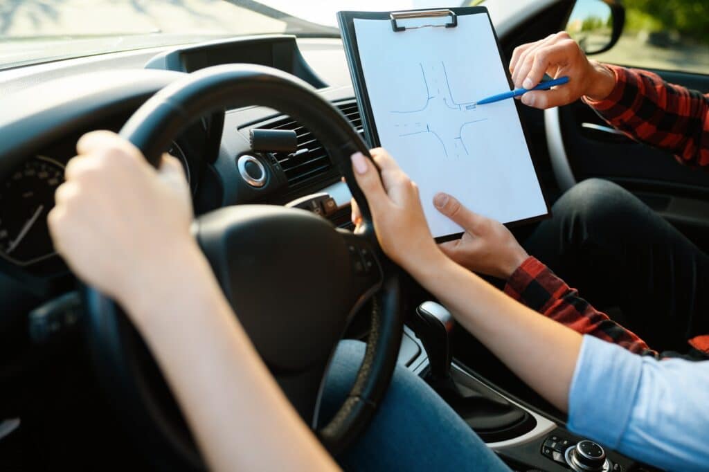 Person in the driver's seat of a car holding the steering wheel, while another person in the passenger seat points at a street diagram on a clipboard, possibly giving driving instructions or lessons.