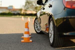 A dark car maneuvers around a line of orange traffic cones on a paved surface, possibly during a driving test or training exercise. The background is slightly blurred, with some greenery and buildings visible in the distance.