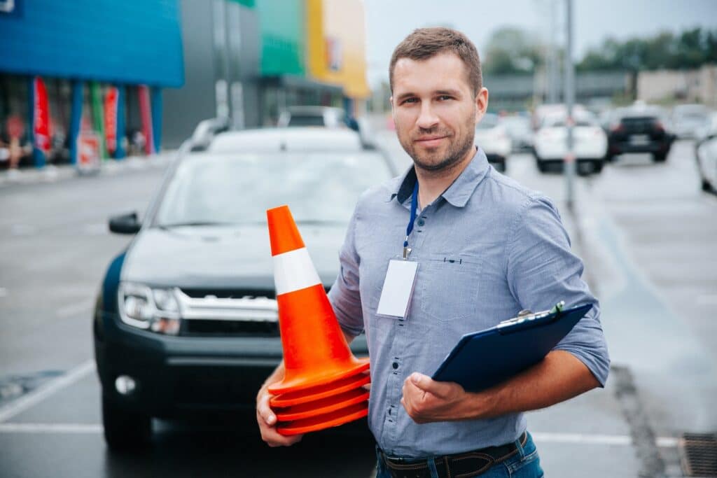 A man stands in a parking lot holding traffic cones and a clipboard. He wears a blue shirt with a blank name tag. Cars are visible in the background, and colorful buildings line the side of the lot. The weather appears overcast.