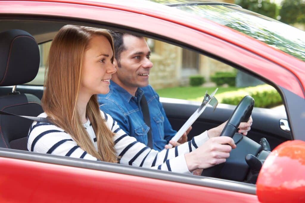 A young woman is sitting in the driver's seat of a red car, smiling while holding the steering wheel. A man with a clipboard is in the passenger seat, observing. They appear to be in a driving lesson or test.