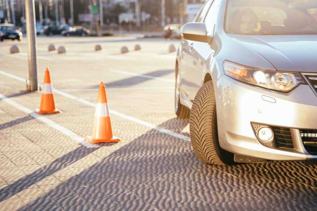 A silver car is parked in a sunlit lot, partially over a marked line. Two orange traffic cones are lined up nearby, casting long shadows on the pavement. The focus is on the car's front tire and side mirror.