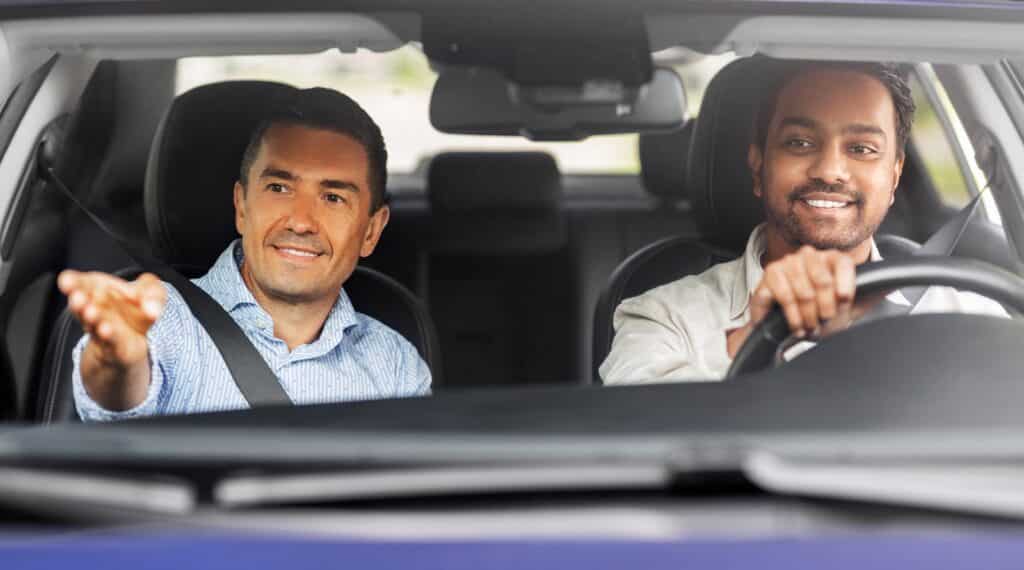 Two men are sitting in the front seats of a car. The man on the left is gesturing while smiling, and the man on the right is driving, also smiling. They appear to be having a conversation while traveling.