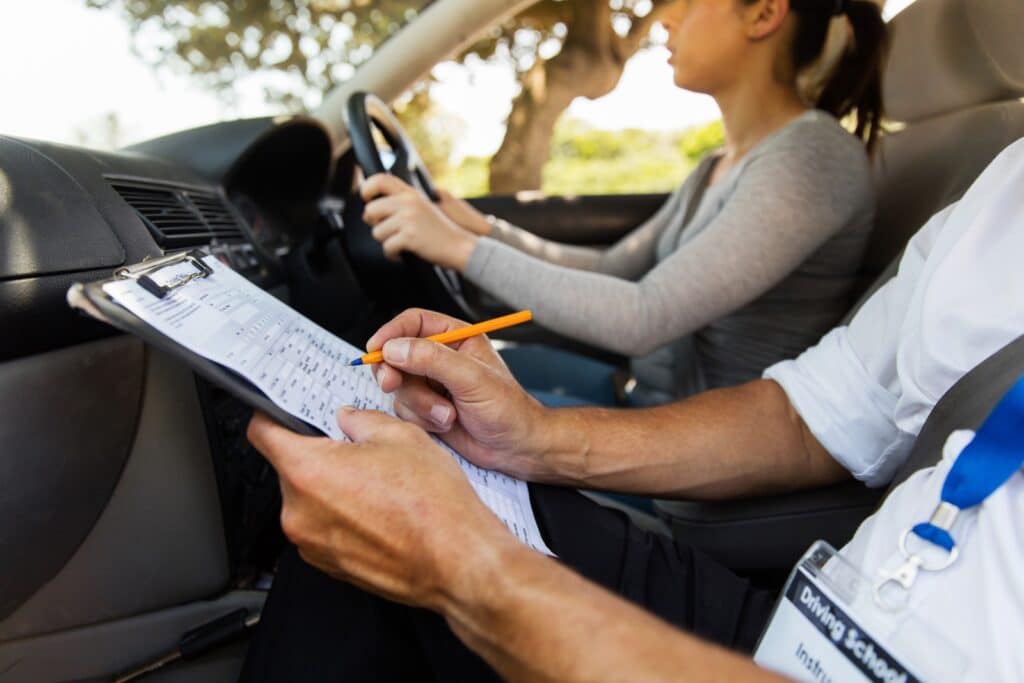 A driving instructor takes notes on a clipboard while sitting in the passenger seat. A young woman is driving, focusing on the road. Sunlight filters through the trees outside the car.