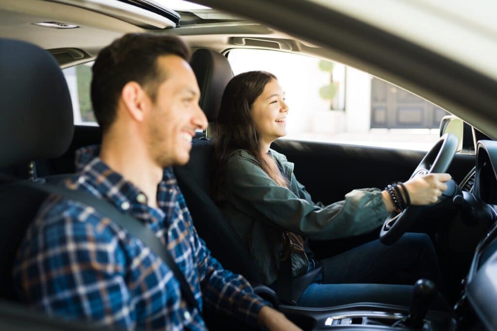 A young woman is smiling as she drives a car, with a man sitting beside her in the passenger seat. Both are wearing seat belts. Sunlight fills the interior of the car, creating a bright and cheerful atmosphere.