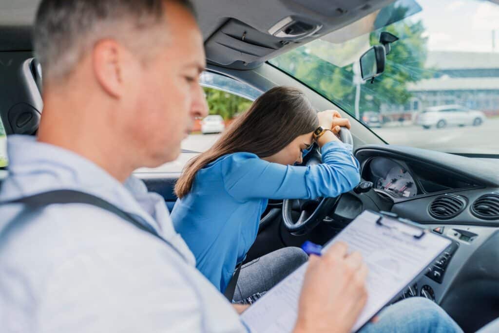 A driving instructor with a clipboard sits beside a frustrated student driver in a car. She's resting her forehead on the steering wheel, expressing stress or disappointment. The street is visible through the windows.