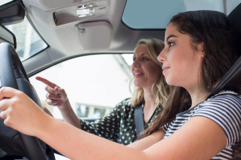 A young woman driving a car while an older woman sits beside her, smiling and pointing ahead. The young woman has both hands on the steering wheel, and they appear to be having a conversation.