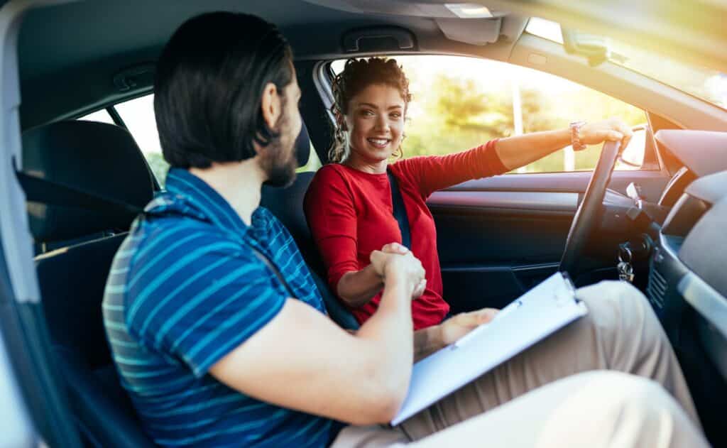 A woman in a red shirt sits in the driver's seat of a car, smiling and shaking hands with a man holding a clipboard in the passenger seat. Sunlight streams in through the window, creating a warm atmosphere.