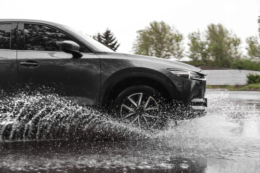 A dark gray SUV is driving through a large puddle on a wet road, causing a dramatic splash of water. Trees and a concrete wall are visible in the blurred background on a cloudy day.