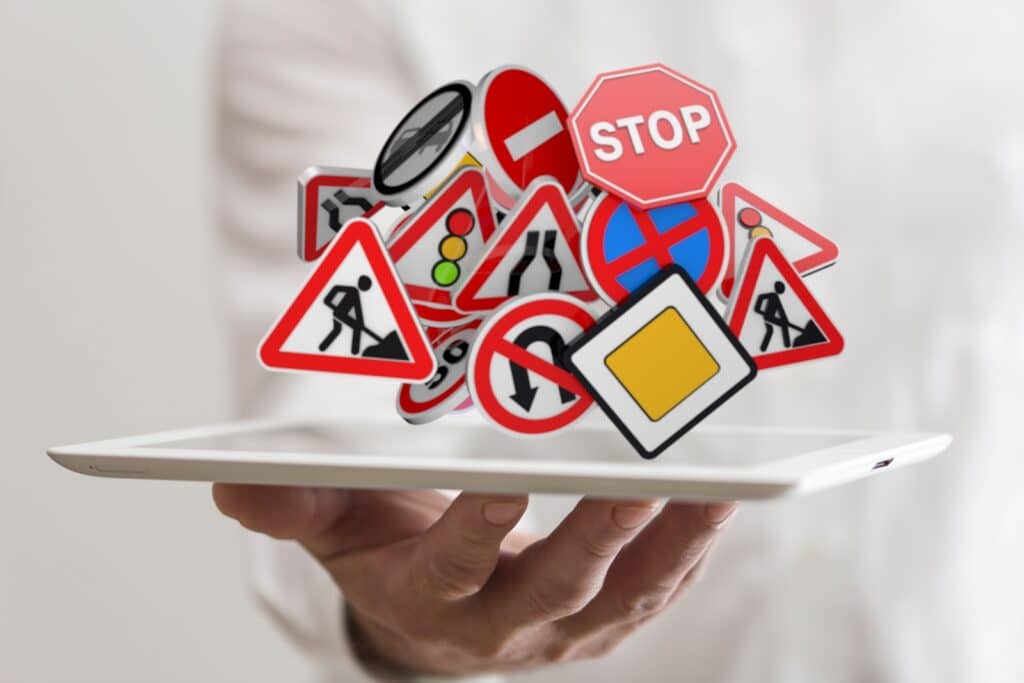 A person holds a tablet with various traffic signs, including stop, pedestrian crossing, and traffic light symbols, floating above it. The background is blurred white, emphasizing the colorful signs.