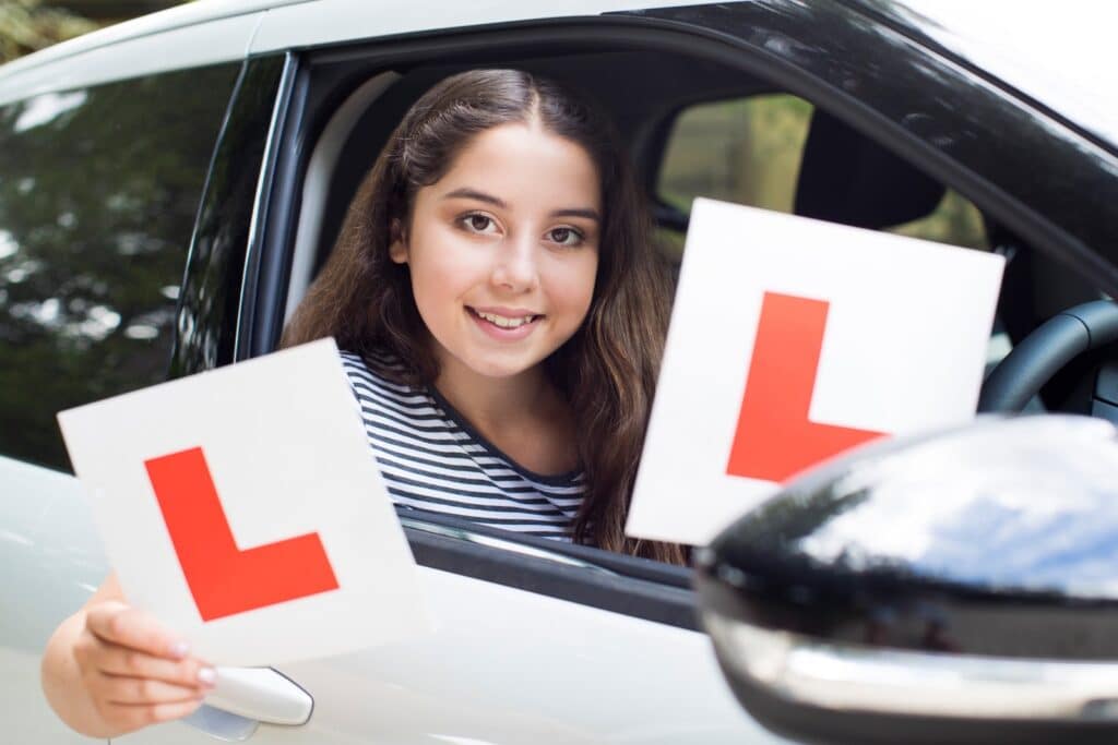 A young woman with long brown hair sits in the driver's seat of a car, smiling, and holds two learner driver signs with red "L" letters out of the window. She wears a striped shirt, and the car is white. The background is blurred greenery.