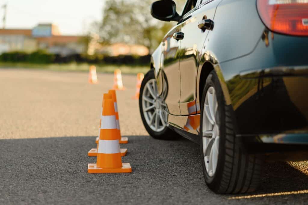 A black car navigates through a slalom course marked by bright orange traffic cones on a sunlit asphalt track. Trees and a building are blurred in the background.