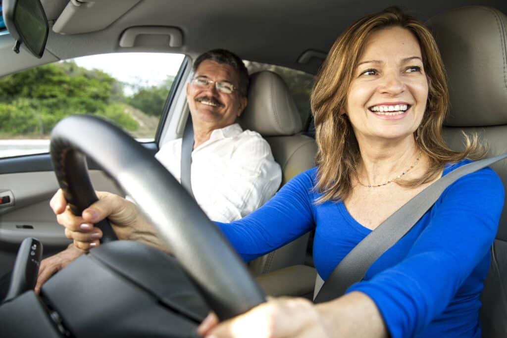 A woman in a blue shirt is happily driving a car, smiling as she holds the steering wheel. A man in a white shirt sits in the passenger seat, also smiling. They appear to be enjoying their drive on a sunny day.