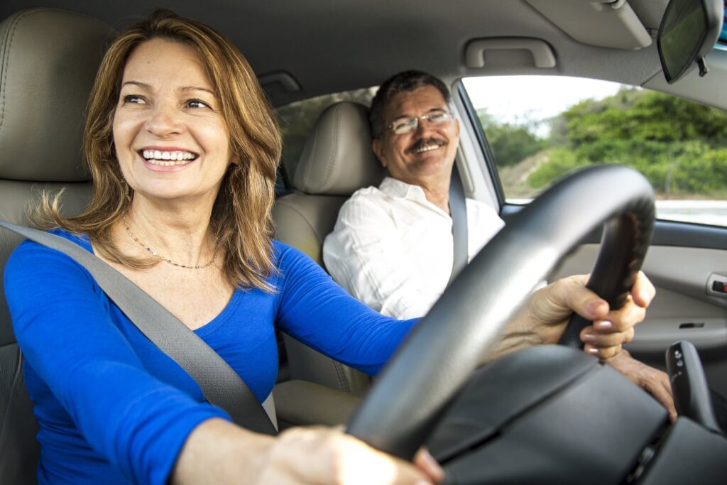 A woman in a blue shirt is happily driving a car, smiling as she holds the steering wheel. A man in a white shirt sits in the passenger seat, also smiling. They appear to be enjoying their drive on a sunny day.