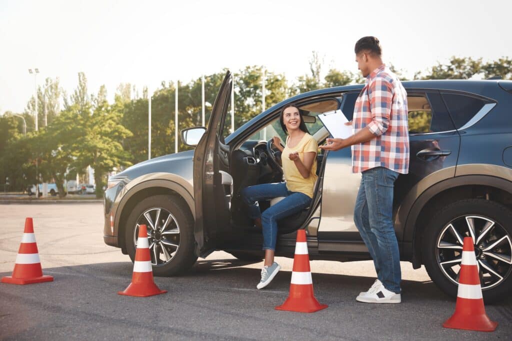 A woman sits in a car with an open door, smiling and giving a thumbs-up. A man stands outside, holding a clipboard. Traffic cones are arranged nearby, suggesting a driving lesson or test setting.