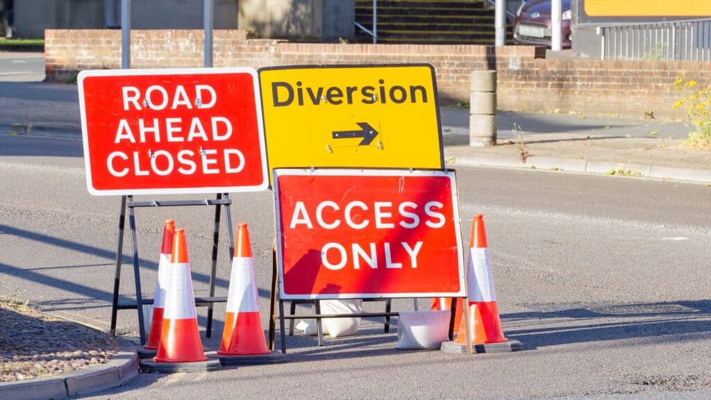 A road junction with three signs: "ROAD AHEAD CLOSED," "ACCESS ONLY," and a yellow "Diversion" sign with a right arrow. Several orange and white traffic cones surround the signs.