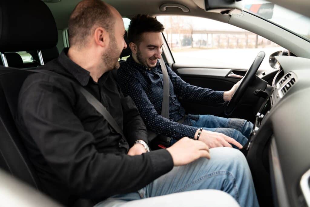 Two men sitting in a car, both wearing seatbelts. The man on the left, in a black shirt, is talking to the man on the right, who is holding the steering wheel and wearing a dark shirt with white dots. They are smiling and engaged in conversation.