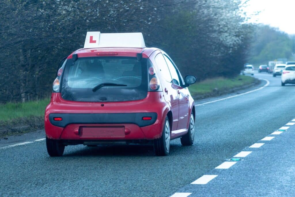 A small red car with an "L" plate on top, indicating a learner driver, travels on a two-lane road surrounded by trees and other vehicles. The sky appears overcast, and the road is slightly wet.