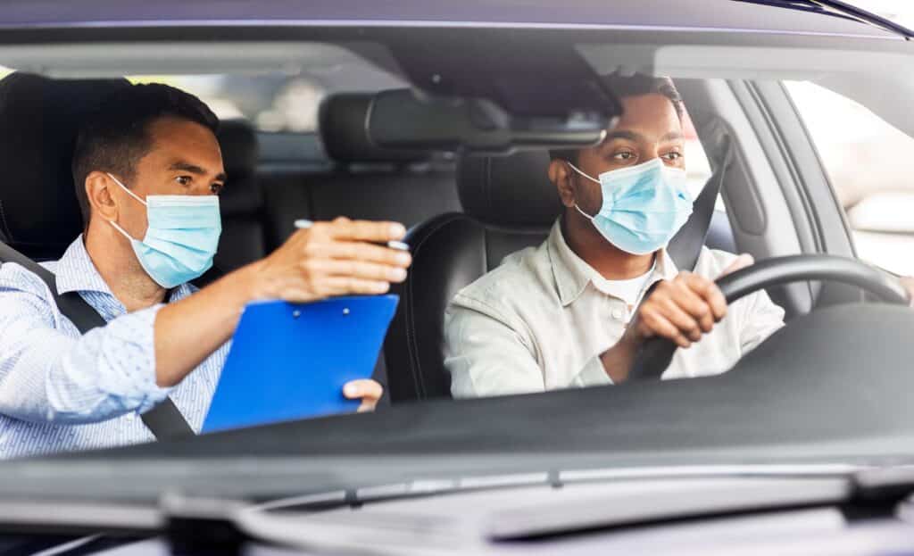 Two people wearing masks are in a car. The person on the right is driving, focused on the road. The person on the left holds a clipboard, pointing ahead, possibly instructing or guiding the driver.