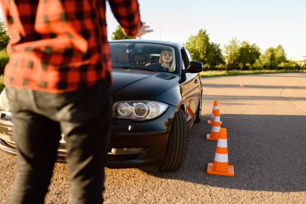 A person in a red plaid shirt is guiding a black car as it maneuvers around orange traffic cones on a sunny day. The driver looks focused. Trees and a clear sky are visible in the background.