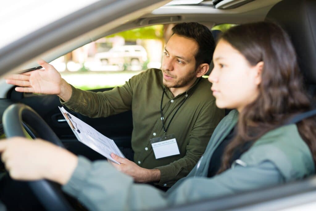 A driving instructor in a green shirt sits in the passenger seat, pointing ahead while holding a clipboard. A young woman in the driver's seat listens attentively, focusing on the road. They are inside a car with blurred greenery outside.