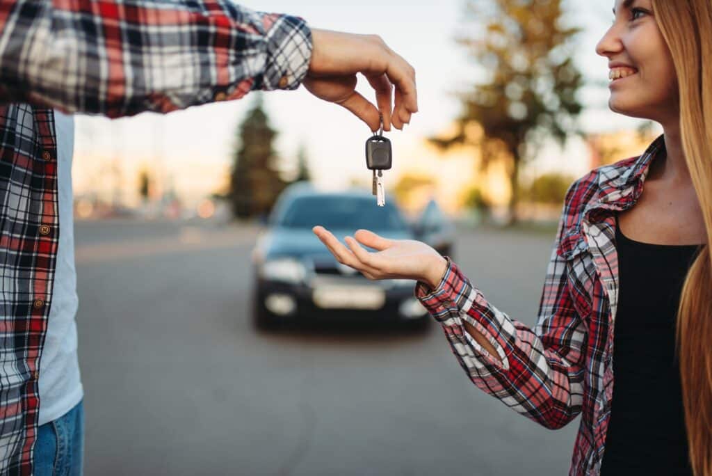 A person in a plaid shirt hands over car keys to a woman with long hair and a plaid shirt. They are standing on a road with a blurred car in the background. The woman is smiling as she reaches for the keys.