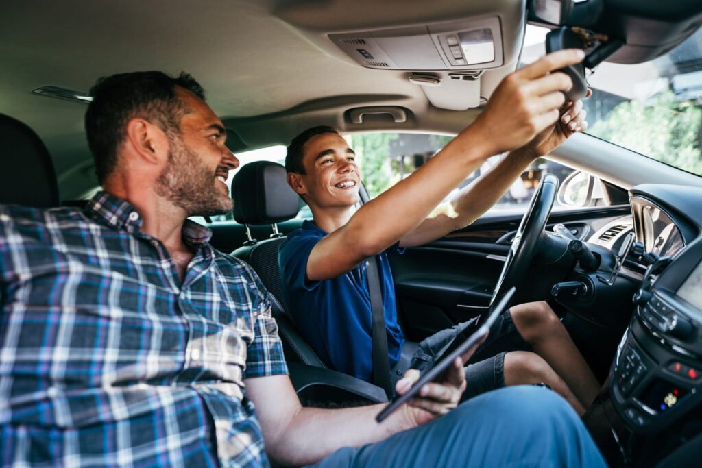 A smiling teen sits in the driver's seat adjusting the rearview mirror, while an adult in the passenger seat looks on approvingly. They are in a parked car with visible sunlight outside.