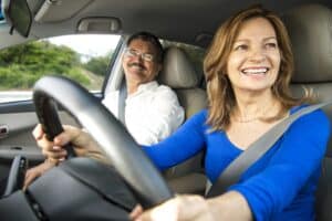 A woman in a blue shirt is driving a car, smiling, with a man as a passenger, also smiling. They are both wearing seatbelts, and the car appears to be in motion. The background shows a blurry view of greenery outside the window.