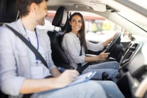 A woman in a striped shirt is smiling while driving a car. She is accompanied by a man in a blue lanyard and light-colored shirt, taking notes on a clipboard. Both are wearing seatbelts and appear to be having a conversation inside the vehicle.