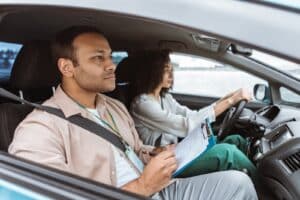 A driving instructor with a notepad sits in the passenger seat while a woman drives a car. Both are focused, and the dashboard is visible. The interior is well-lit, suggesting it's daytime.