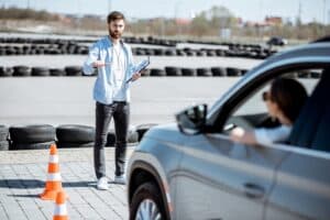 A man stands next to orange traffic cones, holding a clipboard, gesturing to a person in a car during a driving lesson in an outdoor area. A row of tires lines the background, with blurred buildings visible in the distance.