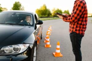 A driving instructor holds a clipboard while standing beside a parked black car with a learner inside. Traffic cones are lined up on the road, and trees are visible in the background.