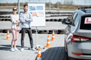 Two people stand smiling beside a car at a driving school. They are outdoors near a flip chart with road signs. Orange traffic cones are arranged around the area, indicating a learning environment.