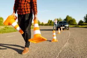 A person in a checked shirt holds traffic cones and walks on a road. More cones are arranged on the ground. In the background, there is a black car parked and a blurred figure standing next to it. Trees line the road under a clear blue sky.