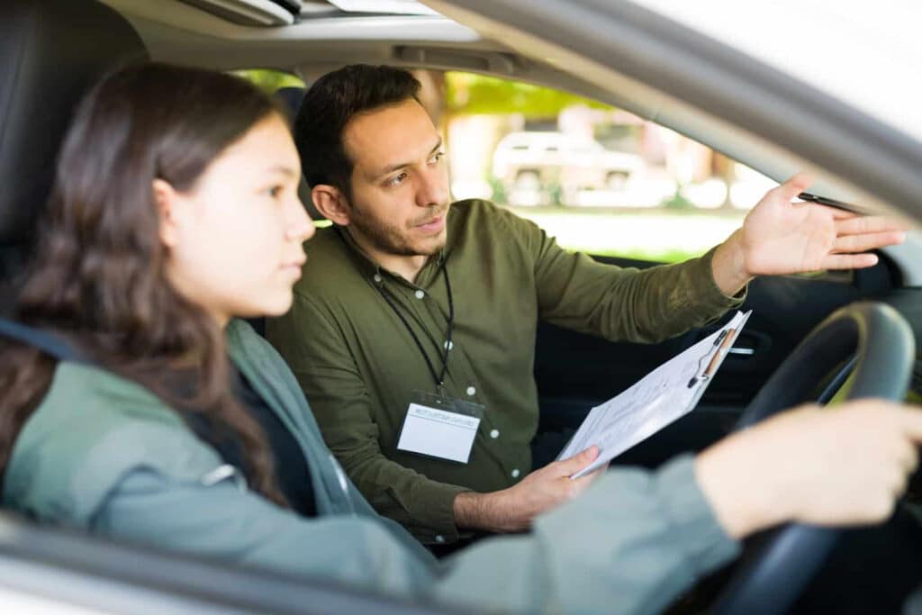 A driving instructor, with a name badge and clipboard, gestures and gives instructions to a young female learner sitting in the driver's seat of a car. They are parked, and the focus is on teaching driving skills.