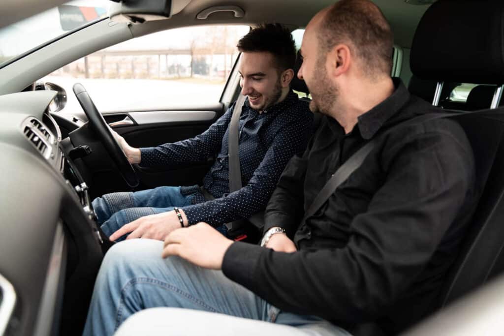 Two men sitting in a car, both smiling. In the driver's seat, one adjusts the controls during an automatic driving lesson, while the bald man observes and gestures towards the dashboard. The bright interior reveals a steering wheel on the right, capturing their shared enthusiasm in Bristol.