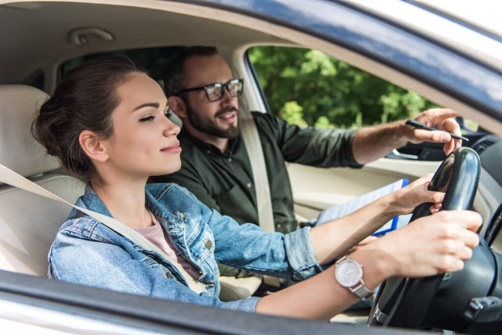 A young woman is driving a car, focusing on the road ahead. A man sitting in the passenger seat is gesturing with his hand, possibly giving instructions. Both are wearing seatbelts. The background shows green foliage outside the car.