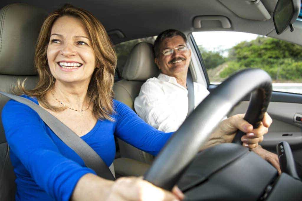 A woman smiles while driving a car, with a male passenger seated next to her. They both appear happy and relaxed. The car interior is visible, and there is greenery outside the window.