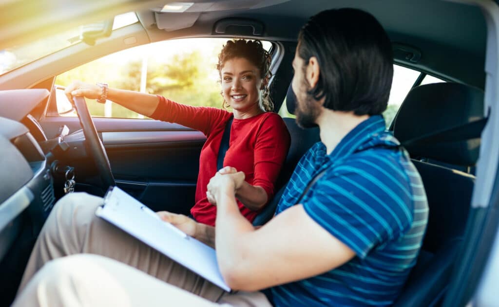 A woman in a red shirt smiles while sitting in the driver's seat of a car, shaking hands with a man with long hair holding a clipboard in the passenger seat. Sunlight filters through the window, creating a warm atmosphere.