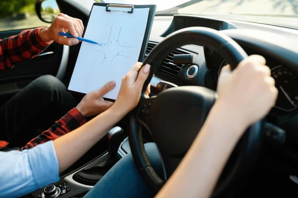 A person in the driver's seat holds the steering wheel while another person next to them points at a road diagram on a clipboard. The view is inside a car, suggesting a driving lesson or instruction.