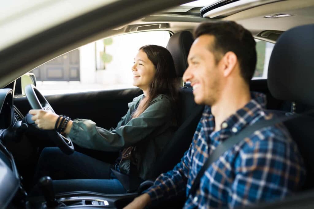 A young woman is driving a car, smiling, with a young man sitting beside her in the passenger seat. Both appear relaxed and happy, enjoying the drive on a sunny day. The car's interior is visible, including the steering wheel and dashboard.