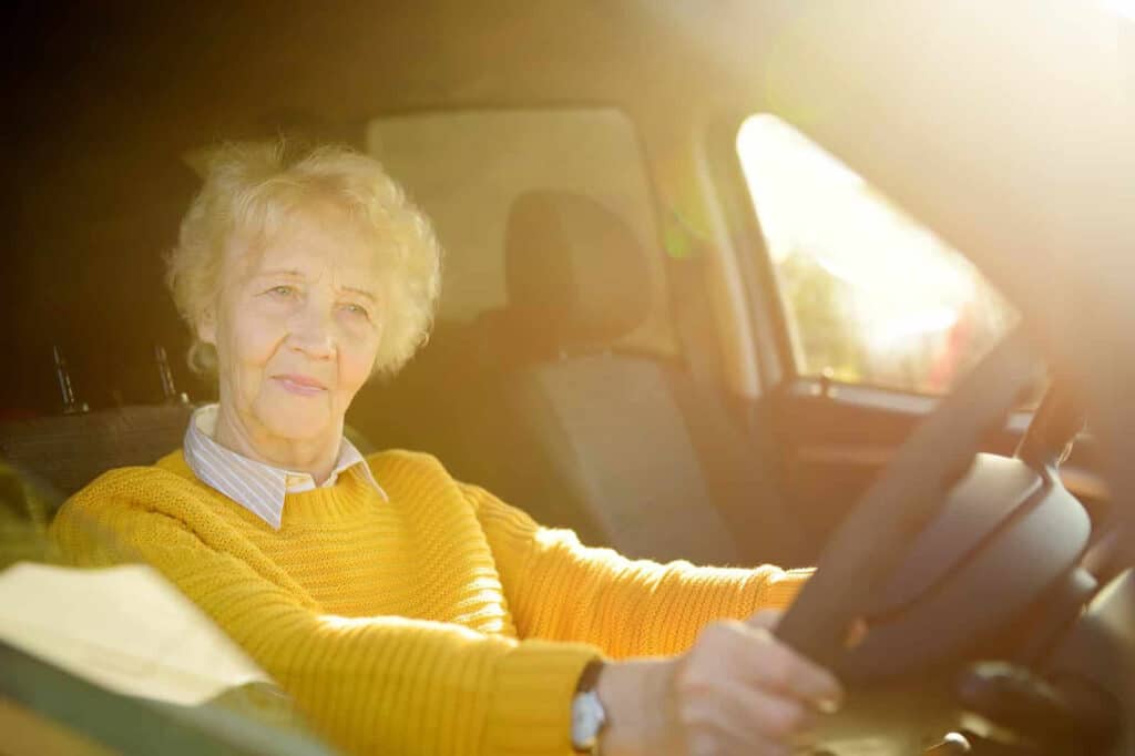 An elderly woman with short, gray hair is sitting in the driver's seat of a car, holding the steering wheel. She is wearing a yellow sweater and a collared shirt. Sunlight is softly illuminating the interior of the car.